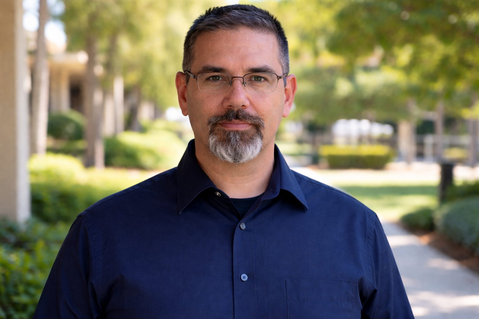 Frank Jamison standing upright and facing the camera in a navy button up shirt and glasses on a sunlit college campus walkway, with trees and buildings softly blurred in the background.