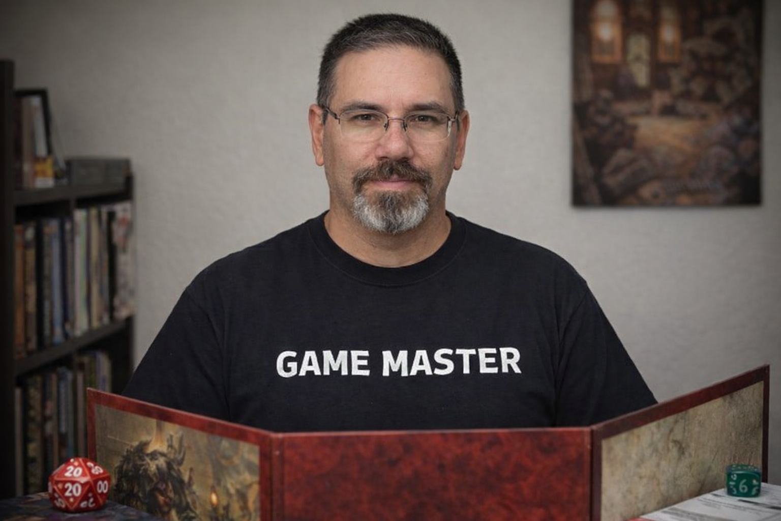 Portrait of Frank seated at a tabletop role playing game setup, facing the camera with a neutral expression. He is wearing glasses and a black shirt labeled Game Master. In front of him is a red dungeon master screen with fantasy artwork, along with polyhedral dice on the table. A bookshelf and framed artwork are softly visible in the background.