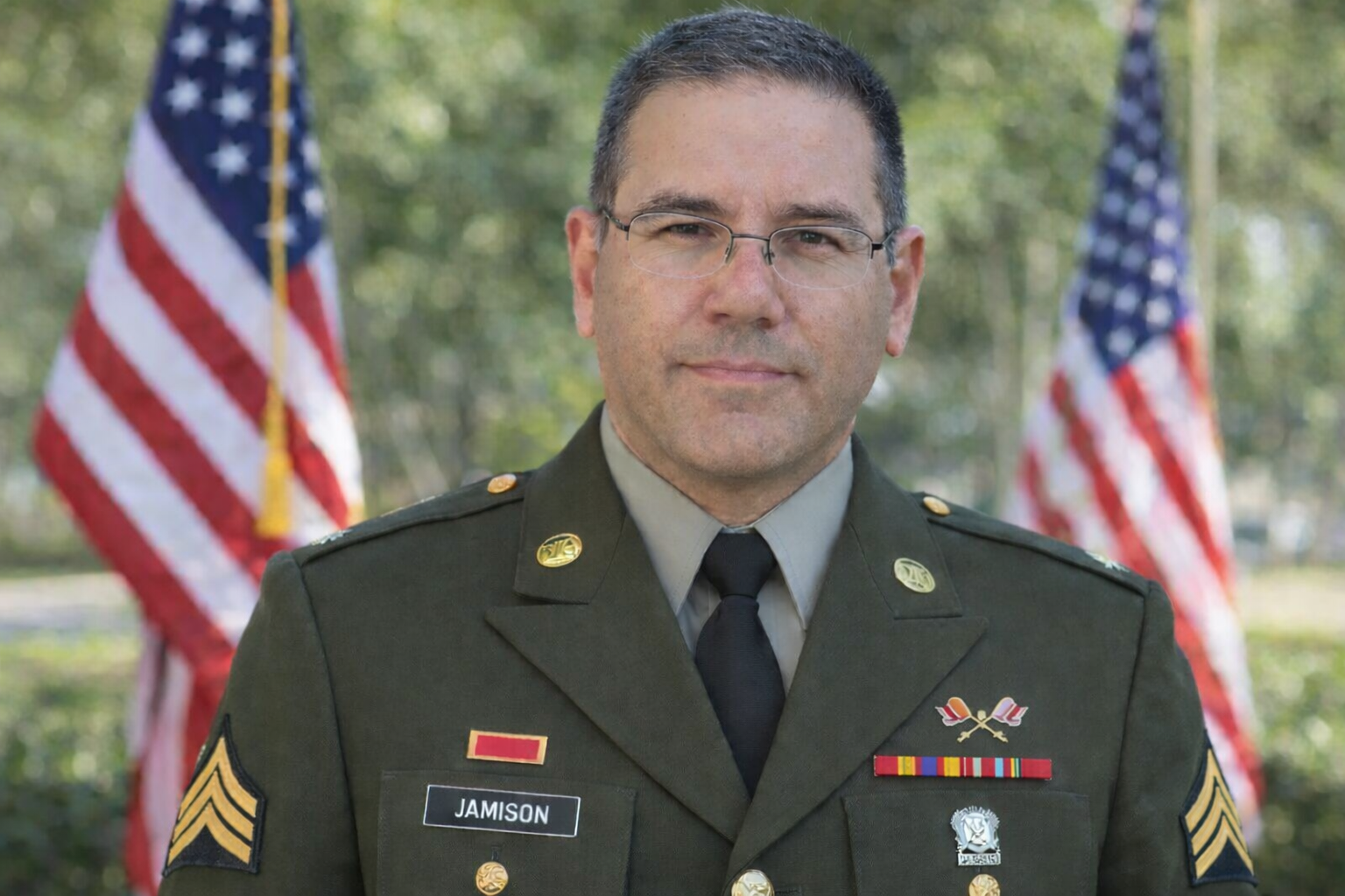 Frank Jamison wearing a U.S. Army green service uniform with sergeant chevrons on both sleeves, a National Defense Service ribbon, and additional service ribbons on his chest. He faces the camera with a composed expression, wearing glasses and a neatly trimmed salt and pepper goatee, standing outdoors with American flags softly blurred in the background.