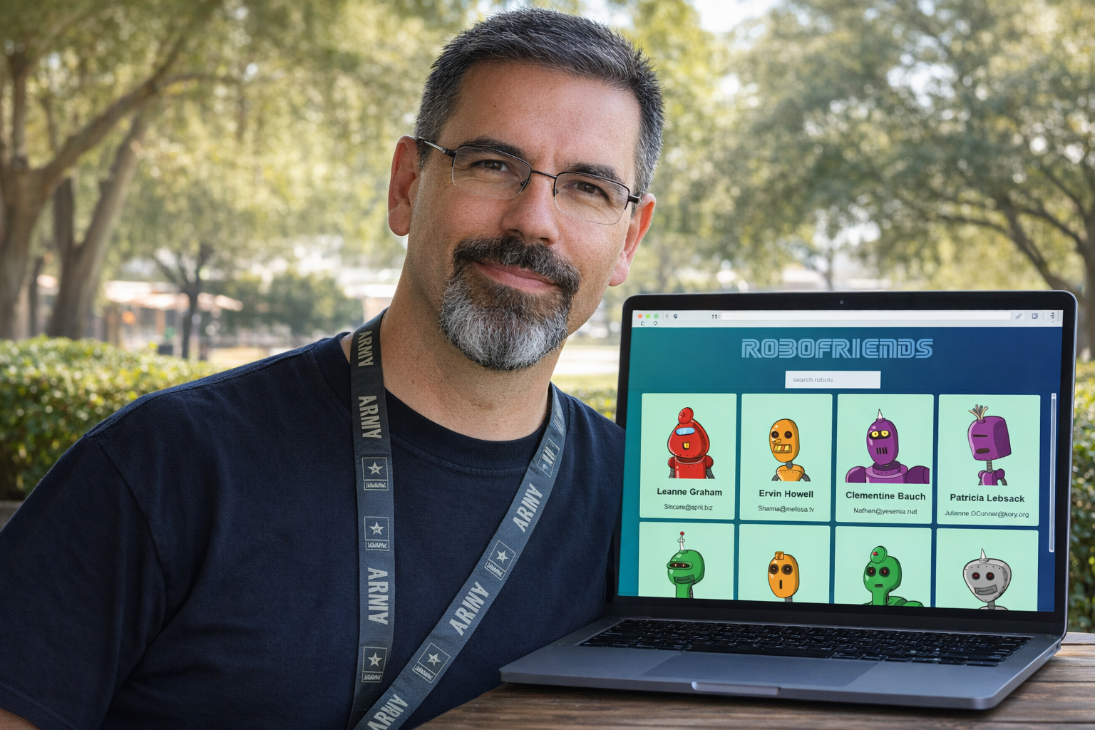 Frank Jamison stands outdoors at a wooden table, facing the camera with a relaxed, confident expression. He is wearing a dark shirt and an Army lanyard. In front of him is an open laptop displaying the RoboFriends web application, featuring a blue gradient background, a ROBOFRIENDS title, a search bar, and a grid of colorful robot avatar cards with names and email addresses. Trees and soft daylight are visible in the background.